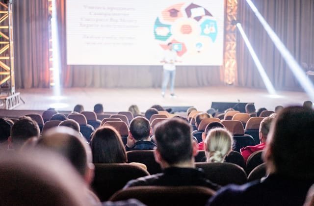 Auditório lotado durante uma palestra, com o palco ao fundo